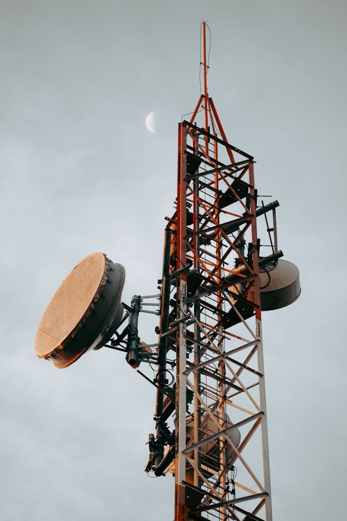 Vertical shot of a telecommunication tower with dishes under a cloudy sky and visible moon.